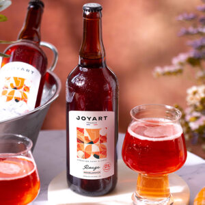 Photo d'ambiance de notre référence houblonnée rouge. L'arrière-plan contient un bouquet de fleur au soleil. Une bouteille est posé sur une table en marbre avec deux verres remplies et derrière une autre bouteille posé dans un seau à glaçon.