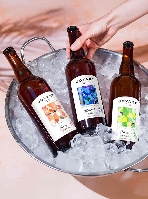 Photo des trois bouteilles de la gamme houblonnée, posé dans un seau à glaçons. À gauche la rouge, au milieu la blanche tenue par une main, et à droite la ginger.