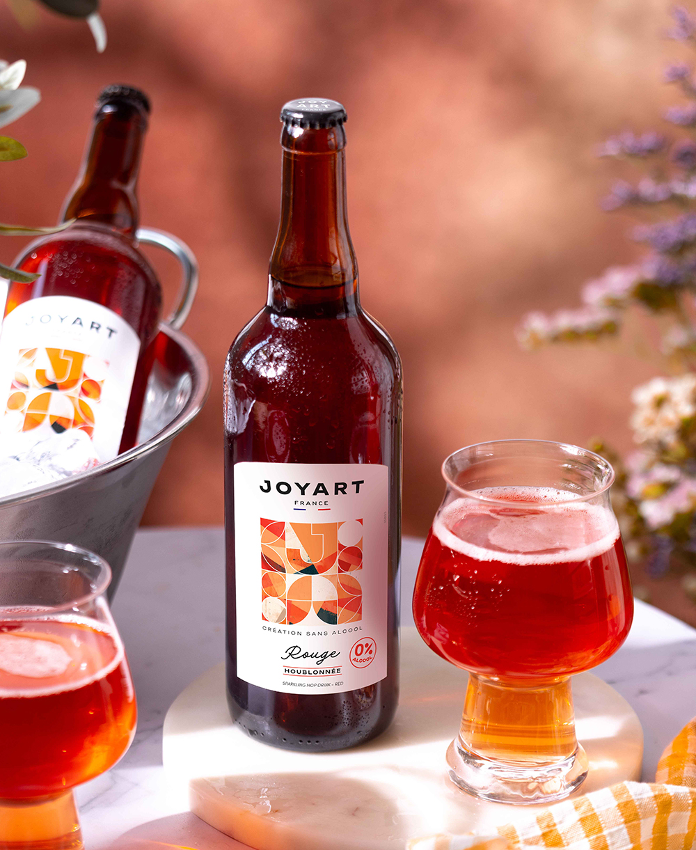 Photo d'ambiance de notre référence houblonnée rouge. L'arrière-plan contient un bouquet de fleur au soleil. Une bouteille est posé sur une table en marbre avec deux verres remplies et derrière une autre bouteille posé dans un seau à glaçon.